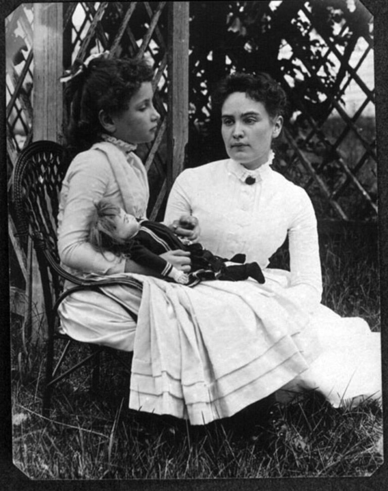 Helen Keller at about the age of seven.  Her hair is curled and pulled back with a ribbon.  She sits with a doll in her lap.  Both her and her teacher wear white dresses.